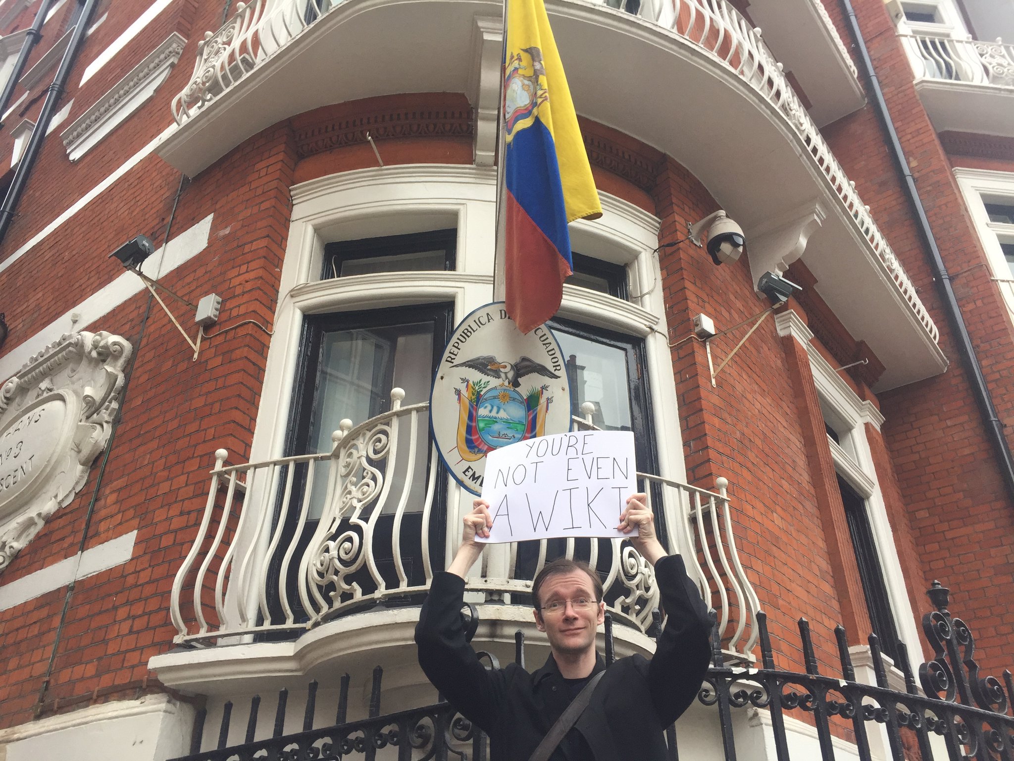 Garrett in 2016, standing outside the Ecuadorian Embassy in London holding a sign saying 