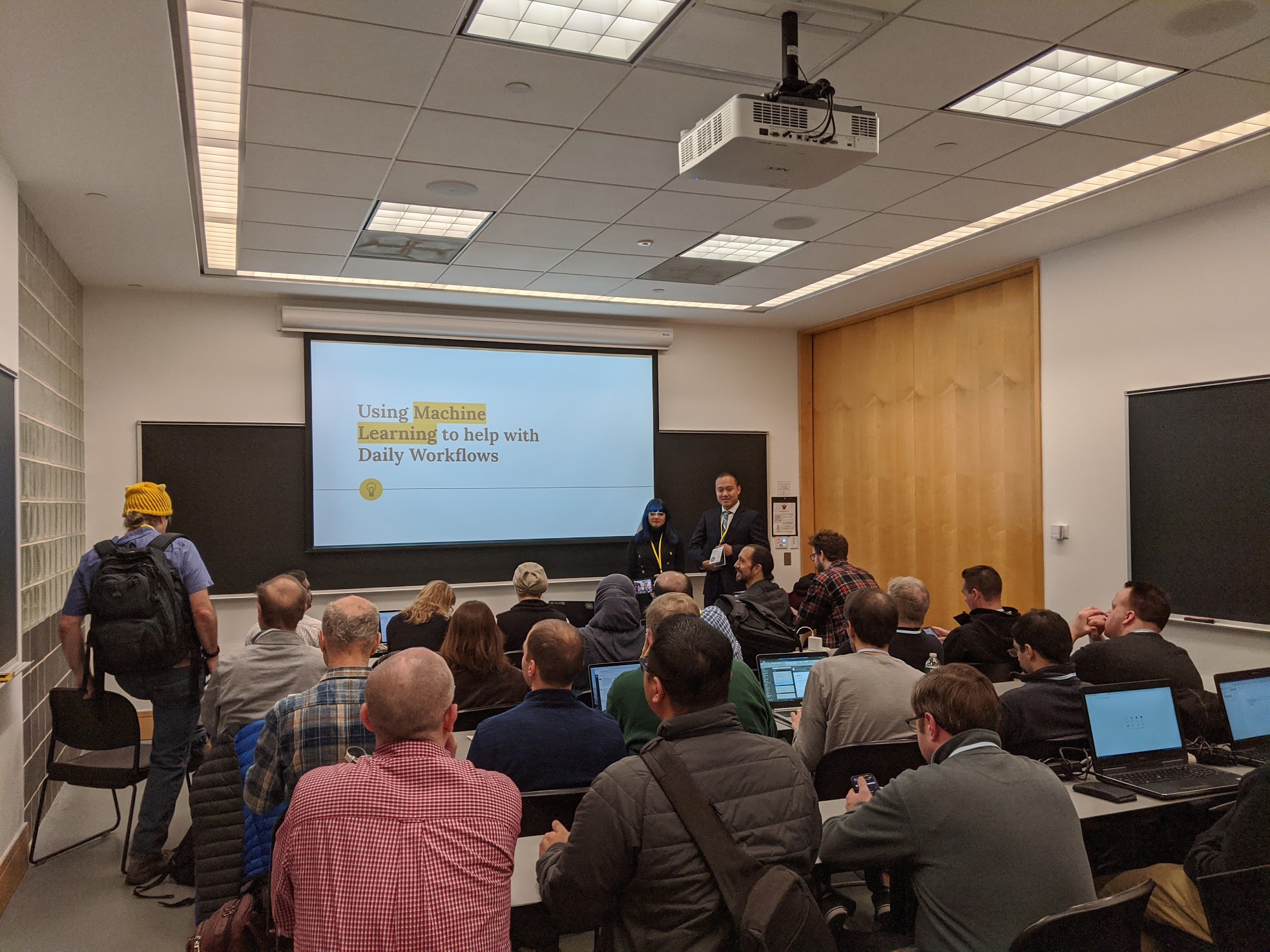 A man and a woman in front of a classroom full of people, the screen behind them says: Using Machine Learning to help with Daily Workflows